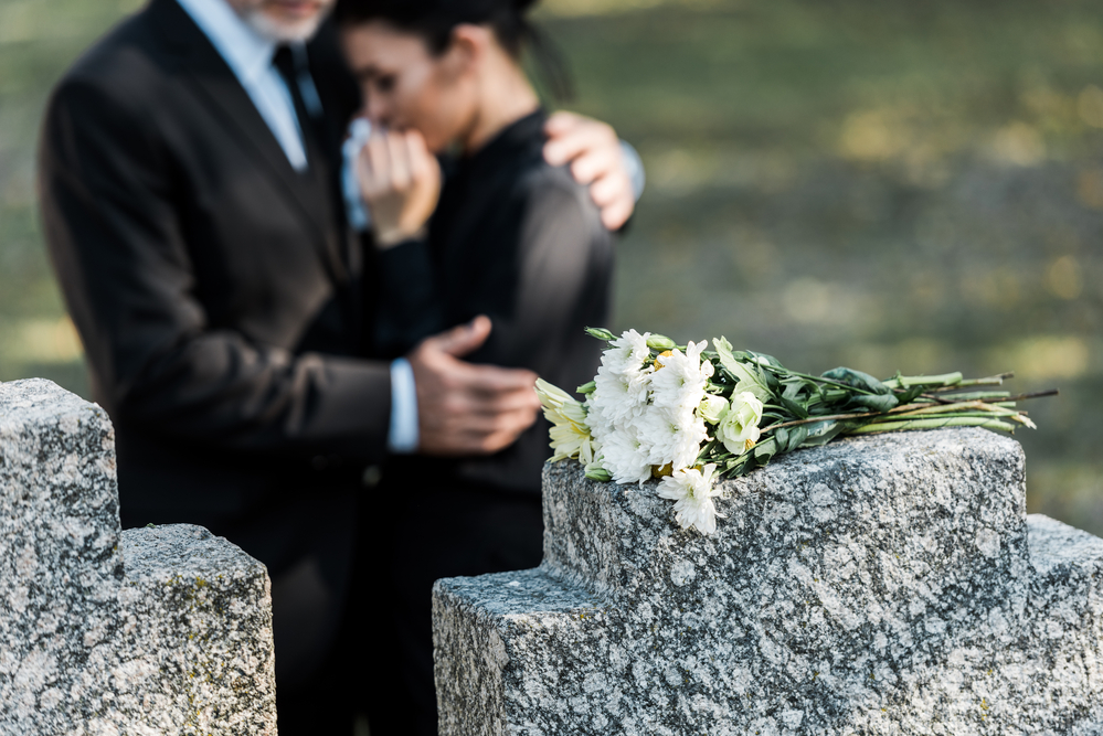 Flowers on tombstone man comforting woman in background
